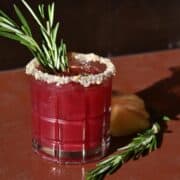 A side view of a cranberry and rosemary mocktail on a table with a sugar rim and rosemary garnish.
