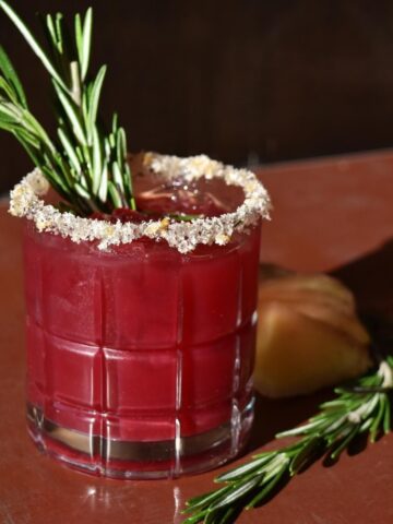 A side view of a cranberry and rosemary mocktail on a table with a sugar rim and rosemary garnish.