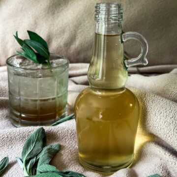 Sage Simple Syrup in a bottle with a drink in the background.