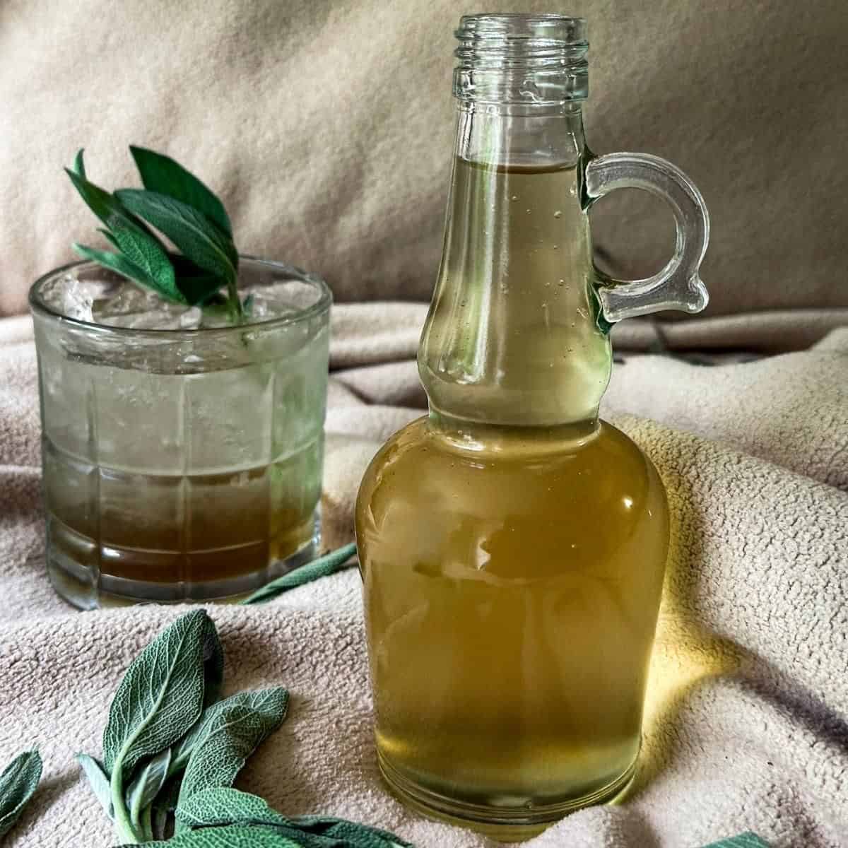 Sage Simple Syrup in a bottle with a drink in the background.