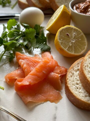smoked salmon, bread, lemon slices, cilantro, and an egg on a white surface