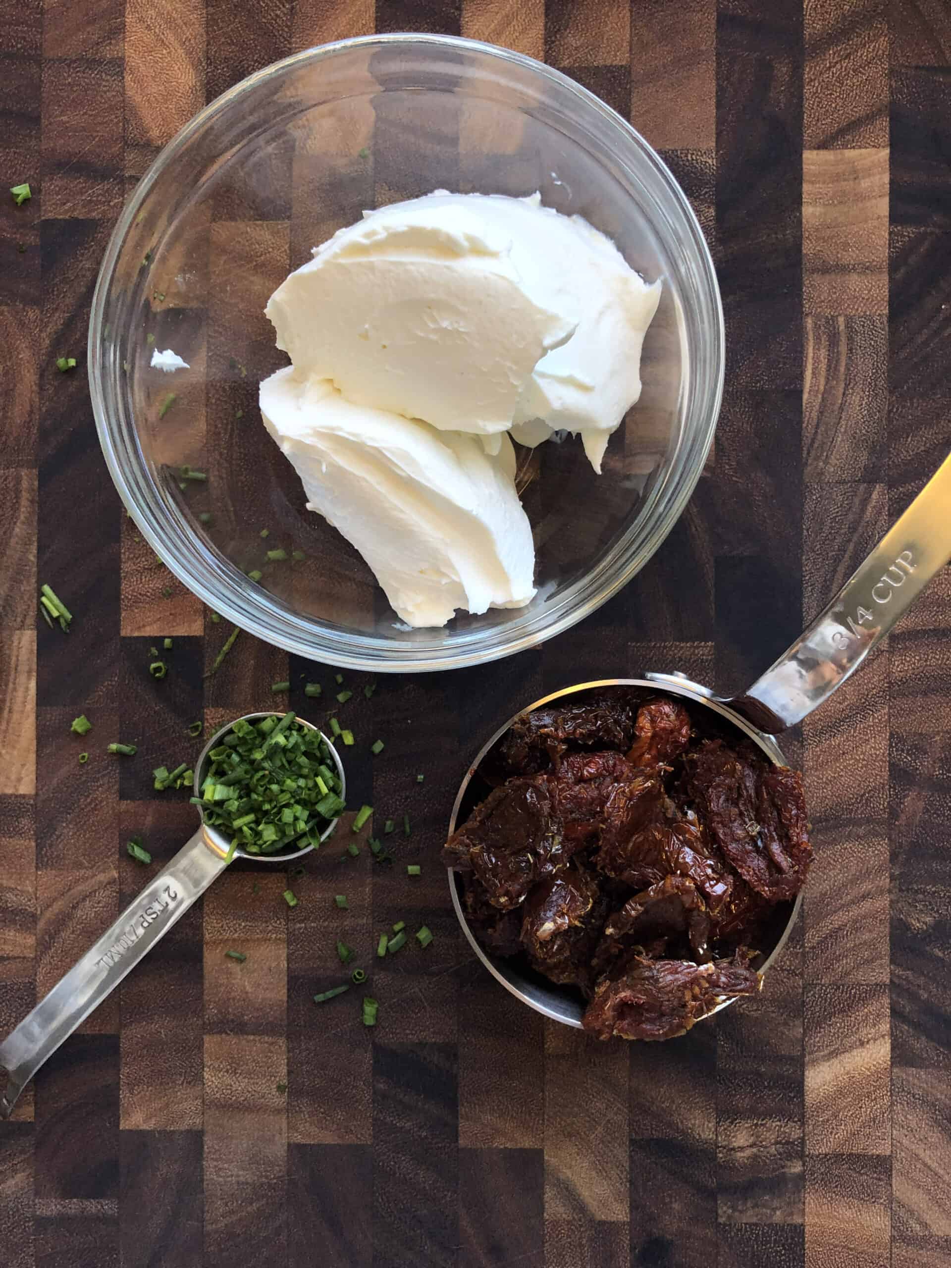 Cream cheese, sun-dried tomatoes, and chopped chives on a cutting board.