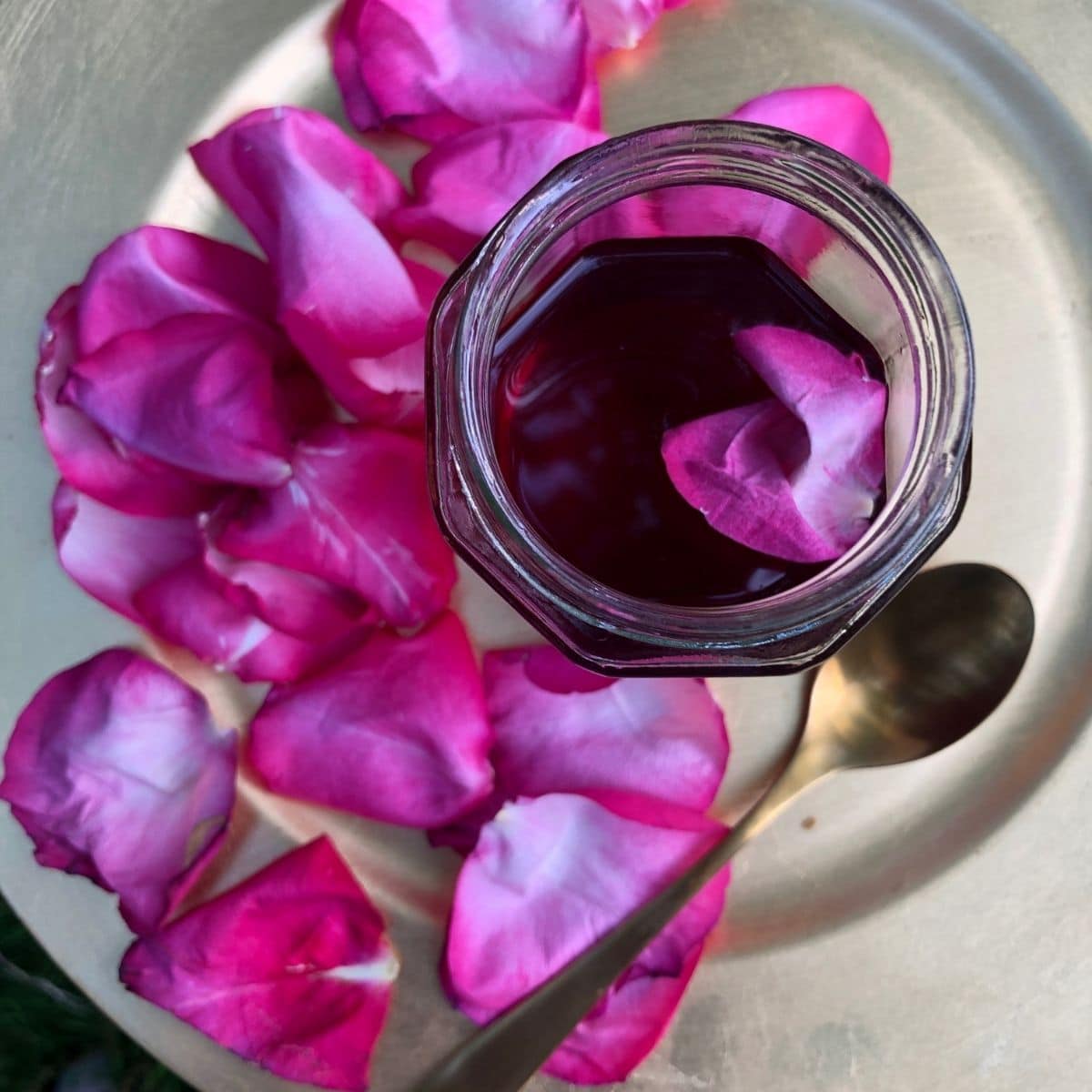An overhead view of rose syrup in a glass jar, on a gold plate with rose petals.