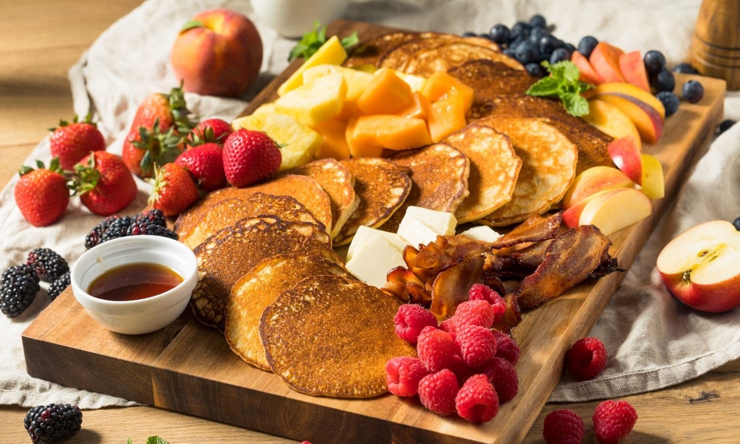 Pancakes, fruit, bacon, and berries on a wood board.
