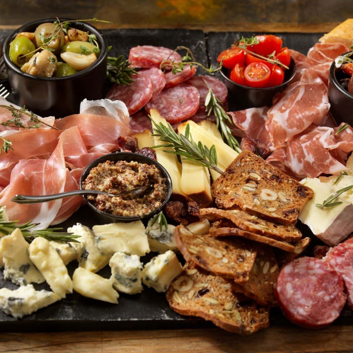 A slate charcuterie board with meats, cheese, and garnishes.